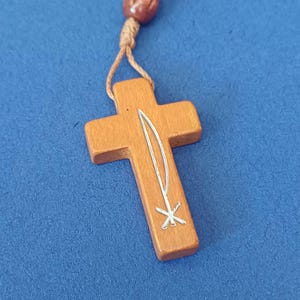 Pu&ograve; includere: Primo piano di un ciondolo a forma di croce in legno con un disegno argentato di una spada e una stella. La croce &egrave; marrone chiaro e lo sfondo &egrave; blu unito. Il ciondolo &egrave; attaccato a un rosario di perline marroni.