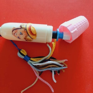 Pu&ograve; includere: Un infila-ago di plastica bianca con un fiocco colorato. L'infila-ago ha una piccola immagine di un bambino con un cappello di paglia. L'infila-ago &egrave; aperto e la parte superiore &egrave; rimossa.
