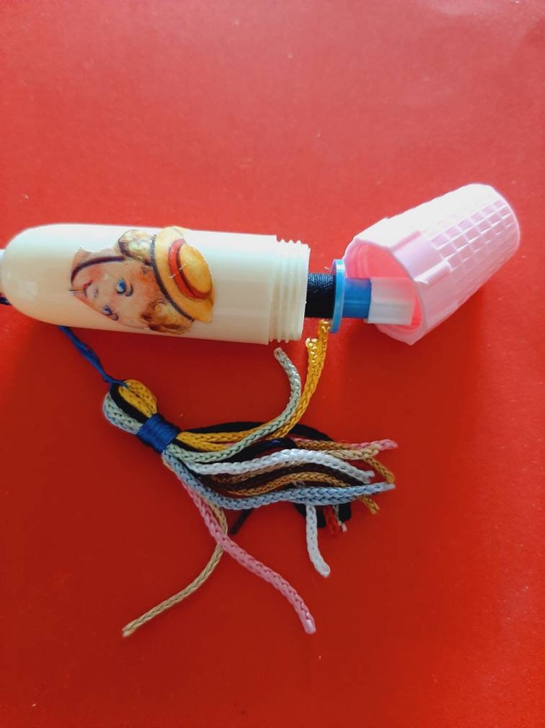 Pu&ograve; includere: Un infila-ago di plastica bianca con un fiocco colorato. L'infila-ago ha una piccola immagine di un bambino con un cappello di paglia. L'infila-ago &egrave; aperto e la parte superiore &egrave; rimossa.