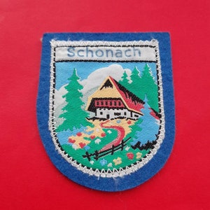 Op de afbeelding: Een blauwe en witte geborduurde patch met het woord "Schonach" in witte letters. De patch toont een klein huis met een rood dak en een kronkelend pad dat er naartoe leidt. Het huis is omgeven door groene bomen en bloemen.