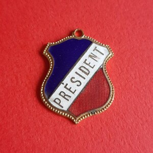 May include: A gold-colored metal shield-shaped charm with a blue, white, and red enamel design. The word "PRESIDENT" is written in white on the charm.