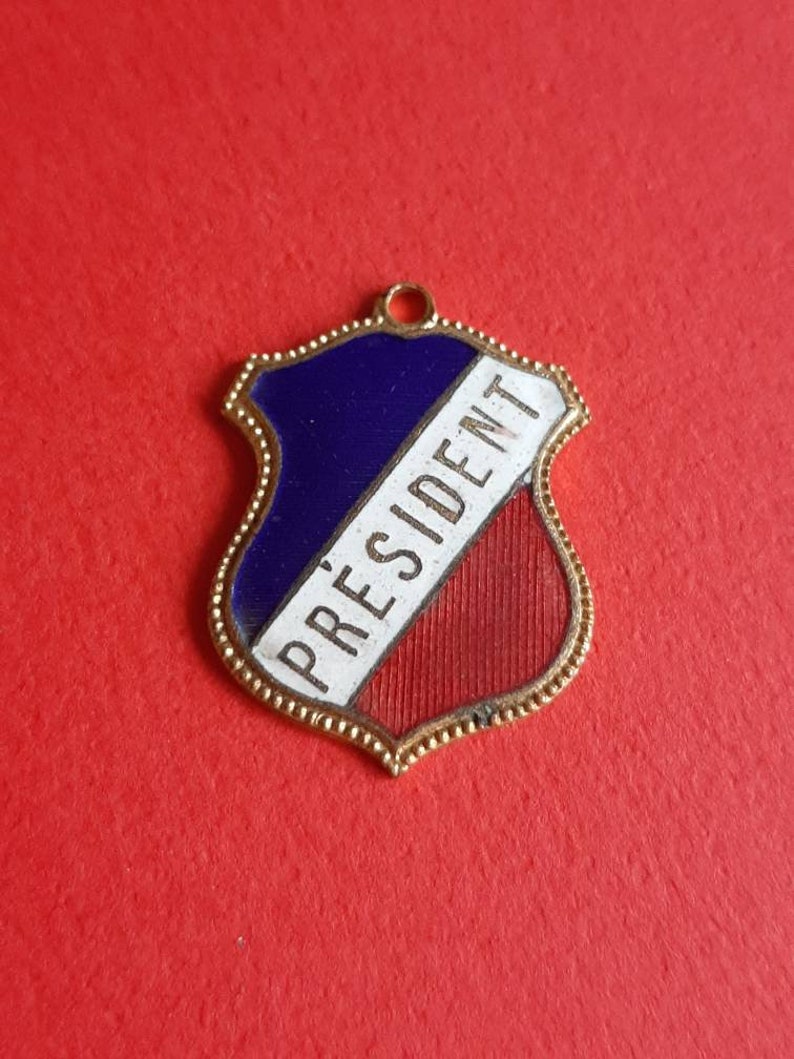 May include: A gold-colored metal shield-shaped charm with a blue, white, and red enamel design. The word "PRESIDENT" is written in white on the charm.