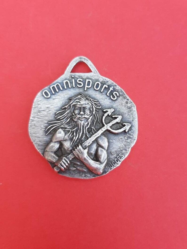 May include: Silver-toned metal keychain with a raised image of a man holding a trident. The keychain is round and has the words "omnisports" and "succes" inscribed on it.