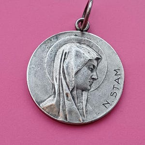 Op de afbeelding: Zilverkleurige hanger met een gedetailleerd reliëf van een vrouwenprofiel, met een sluier en de inscriptie "N. STAM" langs de rand. De hanger heeft een lus aan de bovenkant voor een ketting en staat tegen een roze achtergrond.