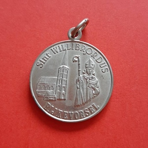 May include: Silver-toned metal pendant featuring a depiction of Saint Willibrordus, a church, and the text "Sint Willibrordus,  Rijkvorsel".