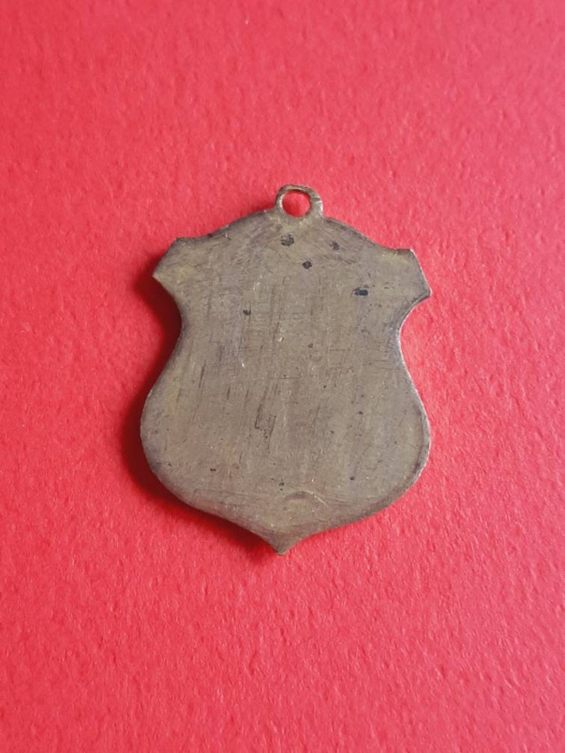 May include: A blank, shield-shaped metal pendant with a loop at the top. The pendant is a light brown color and has a slightly textured surface.