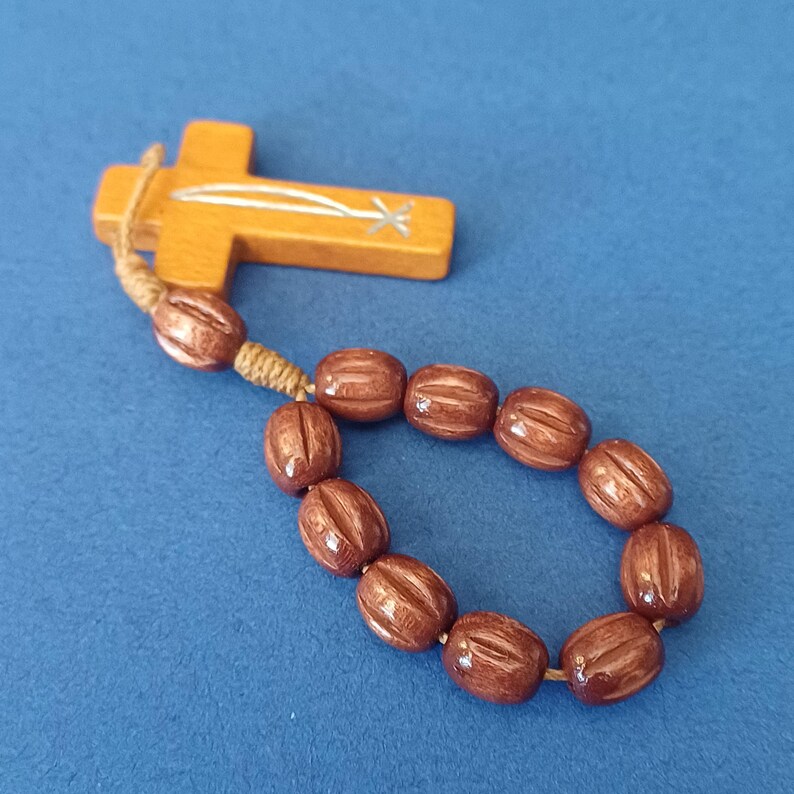 Pu&ograve; includere: Un rosario in legno con grani marroni e una croce. La croce &egrave; marrone chiaro con un disegno argentato. Le perle ovali hanno una finitura marrone scuro lucida. Il rosario &egrave; su una superficie blu.