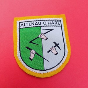 May include: A yellow, green, and white embroidered patch with the text "ALTENAU-O.HARZ" in black. The patch features a stylized design of three crossed mining tools.