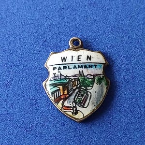 Könnte beinhalten: Ein schildförmiger Anhänger mit goldfarbenem Rand. Der Anhänger zeigt eine gemalte Szene einer Stadt mit dem Text "WIEN PARLAMENT" in Schwarz. Der Anhänger hat oben eine kleine Schlaufe zur Befestigung und ist vor blauem Hintergrund platziert.