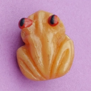 May include: A small, orange, ceramic frog figurine with black and red eyes. The frog is sitting on a purple surface.