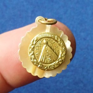 Peut inclure: Un pendentif rond composé d'un médaillon circulaire doré en relief représentant une silhouette drapée, entouré d'une couronne. Le médaillon est fixé sur une base en nacre crème festonnée, surmontée d'un anneau de suspension doré.