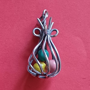 Op de afbeelding: Een zilverkleurige hanger met een kooi-achtig ontwerp, met drie kleurrijke kralen. De kralen zijn groen, geel en roze. De hanger heeft een lus aan de bovenkant voor een ketting, tegen een rode achtergrond.