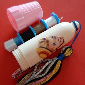 Pu&ograve; includere: Un distributore di filo da cucire in plastica bianca con un fiocco colorato e un'illustrazione vintage di un bambino con un cappello di paglia. Il distributore ha tre bobine di filo, una bianca, una marrone e una nera.