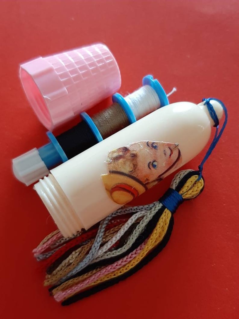 Pu&ograve; includere: Un distributore di filo da cucire in plastica bianca con un fiocco colorato e un'illustrazione vintage di un bambino con un cappello di paglia. Il distributore ha tre bobine di filo, una bianca, una marrone e una nera.
