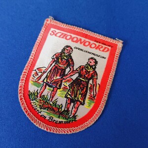 Könnte beinhalten: Ein rechteckiger Aufnäher mit rotem Rand und weißem Innenraum. Der Aufnäher zeigt zwei Figuren in traditioneller Kleidung, die Werkzeuge halten, sowie den Text "SCHOONOORD OPENLUCHTMUSEUM" und "Ellert en Brammert".