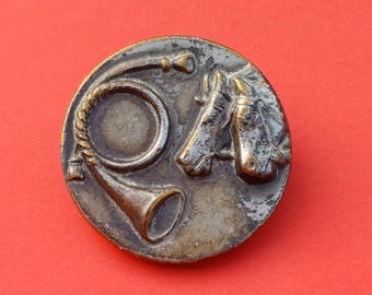 Könnte beinhalten: Ein runder Metallknopf mit einer verwitterten, dunklen Oberfläche. Das Relief zeigt ein kreisförmig gewundenes Jagdhorn und zwei nebeneinander angeordnete Pferdeköpfe in einem metallischen Bronzeton.