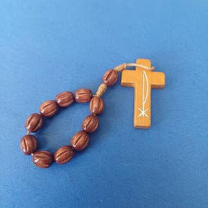 Pu&ograve; includere: Un rosario in legno con perline marroni e una croce in legno. La croce ha un disegno bianco ed &egrave; collegata alle perline con uno spago beige. Il rosario &egrave; posto su uno sfondo blu.