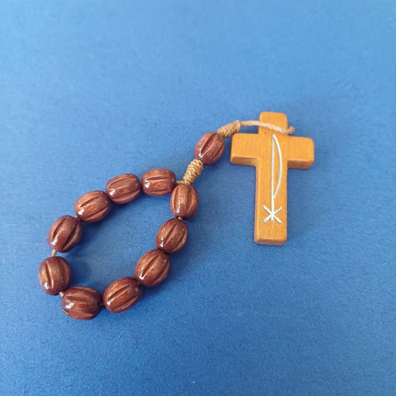 Pu&ograve; includere: Un rosario in legno con perline marroni e una croce in legno. La croce ha un disegno bianco ed &egrave; collegata alle perline con uno spago beige. Il rosario &egrave; posto su uno sfondo blu.