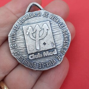May include: Silver-toned metal medallion with the text "SI JE VEUX-LE BONHEUR SI JE VEUX-LE BONHEUR.SI JE VEUX-LE BONHEUR" around the edge. The center of the medallion features a stylized trident with the text "Club Med" below it.