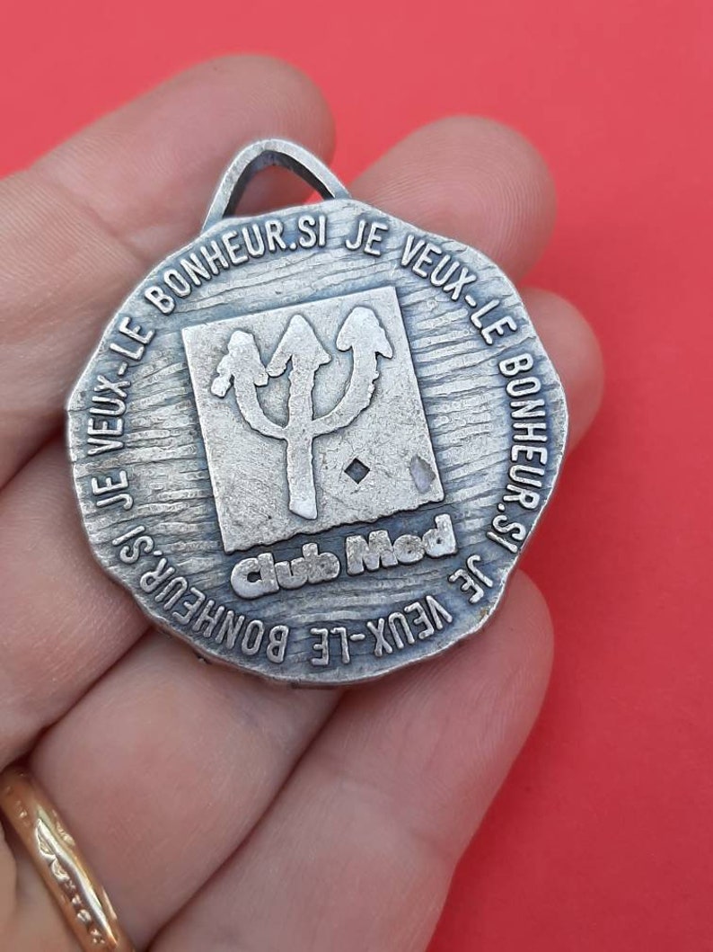 May include: Silver-toned metal medallion with the text "SI JE VEUX-LE BONHEUR SI JE VEUX-LE BONHEUR.SI JE VEUX-LE BONHEUR" around the edge. The center of the medallion features a stylized trident with the text "Club Med" below it.