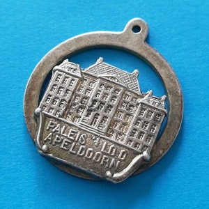 Può includere: Ciondolo in argento con un'immagine dettagliata del Palazzo Het Loo ad Apeldoorn, Paesi Bassi. Il ciondolo è rotondo e ha un foro nella parte superiore per essere attaccato a un braccialetto o una collana.