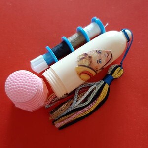 Pu&ograve; includere: Un kit da cucito in plastica bianca con un fiocco colorato, tre bobine di filo e un cuscino per bottoni rosa. Il kit presenta un'illustrazione vintage di un bambino con un cappello di paglia.