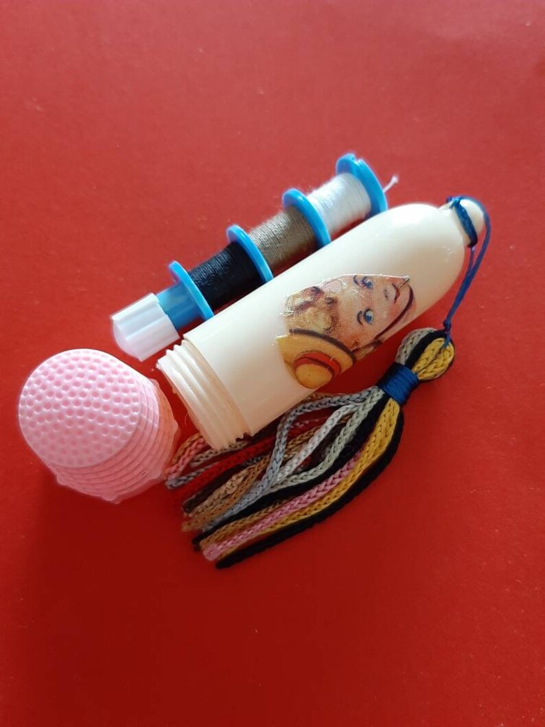 Pu&ograve; includere: Un kit da cucito in plastica bianca con un fiocco colorato, tre bobine di filo e un cuscino per bottoni rosa. Il kit presenta un'illustrazione vintage di un bambino con un cappello di paglia.