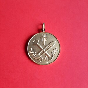 May include: A gold-toned pendant with a cross and two crossed tools, surrounded by a wreath of leaves.