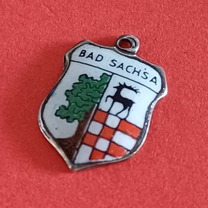 Op de afbeelding: Zilveren bedel in de vorm van een schild met emaille details. Het schild heeft een groene boom, een zwart hert en een rood-wit geruit patroon. De tekst "BAD SACHSÄ" staat in wit bovenaan het schild.