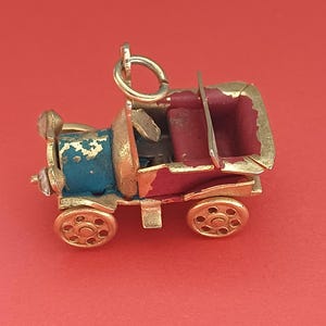 May include: A gold-toned metal charm shaped like a vintage automobile. The car features a blue hood, a deep red interior, and gold-colored wheels. A small circular jump ring is attached to the top of the vehicle for hanging as a jewelry piece.
