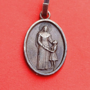 Puede incluir: Pendiente de plata con una imagen en relieve de una mujer con una túnica larga que sostiene la mano de un niño. La mujer lleva una cubierta para la cabeza y el niño lleva un vestido. El pendiente tiene forma ovalada y tiene un bucle en la parte superior para colgar.