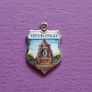 Op de afbeelding: Een zilveren bedel in de vorm van een schild met een blauw, wit en geel emaille ontwerp. De bedel toont een gebouw met de tekst "PORTA WESTFALICA" erboven.