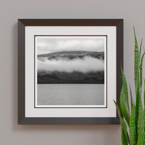 May include: Black and white framed print of a mountain landscape with low-lying clouds over a lake. The print is in a dark brown frame with a white mat. A green plant is to the right of the frame.