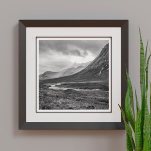 May include: Black and white framed print of a mountain landscape with a river winding through a valley. The artwork is displayed on a gray wall, with a green plant in the foreground.