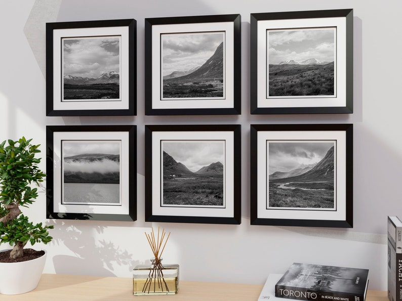 May include: Six framed landscape photographs in black and white, mounted on a white wall. Each photo is encased in a black frame with a white mat. The images depict mountainous terrain and cloudy skies, creating a cohesive gallery wall.