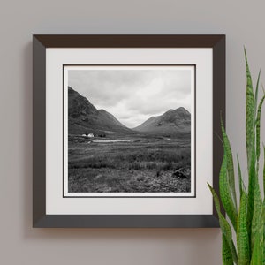 May include: Black and white framed print of a scenic landscape. The photograph captures a valley between two mountains under a cloudy sky. A small building is visible near a body of water in the distance. A green plant is in the foreground.