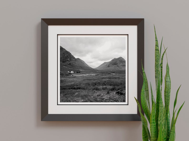 May include: Black and white framed print of a scenic landscape. The photograph captures a valley between two mountains under a cloudy sky. A small building is visible near a body of water in the distance. A green plant is in the foreground.