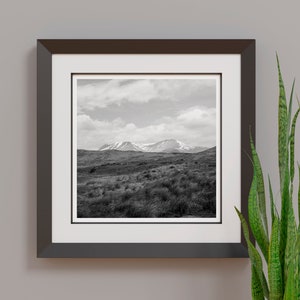 May include: Black and white framed print of a mountain landscape. The artwork features a view of rolling hills and mountains under a cloudy sky. The frame is dark brown, and a green plant is visible on the right.