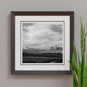 May include: Black and white framed print of a mountain landscape. The photograph features snow-capped mountains under a cloudy sky. The frame is dark brown, and the print is matted with a white border. A green plant is visible on the right.