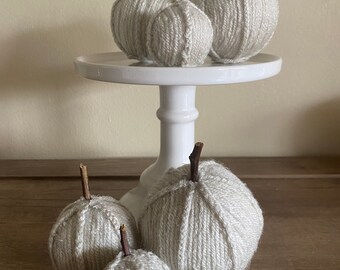 May include: Five yarn pumpkins of varying sizes, all in a light gray color, are displayed on a white pedestal and a wooden surface. The pumpkins have twig stems.