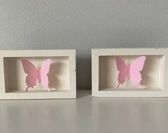 May include: Two white picture frames with pink butterfly cutouts inside. The frames are on a white surface.