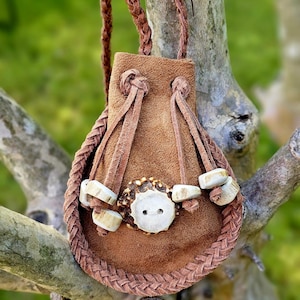 May include: A brown suede leather pouch with a braided trim and a braided strap. The pouch is decorated with antler pieces and a button. The bag is hanging on a tree branch.