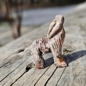 May include: A carved stone sculpture of a wolf in a standing pose. The sculpture is primarily beige with reddish-brown and orange accents. The wolf is standing on a weathered wooden surface, with a blurred background.