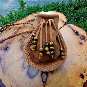 May include: A brown suede pouch with a braided leather trim and drawstring closure. The pouch is adorned with beads and a long braided strap. The pouch is displayed on a wooden surface.