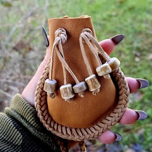 May include: A small, brown leather pouch with a braided border and drawstring closure. The pouch features decorative wooden beads and is held in a hand with long, dark fingernails. The background is blurred, suggesting an outdoor setting.