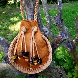May include: A tan leather drawstring pouch featuring a braided leather border and a braided leather shoulder strap. The drawstring closure is accented with four dangling leather cords, each finished with a faceted tiger eye gemstone bead.