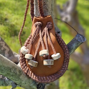 May include: A brown leather pouch with a braided border and drawstring closure. The pouch has a long braided strap and is adorned with wooden beads. The bag is hanging from a tree branch against a blurred green background.