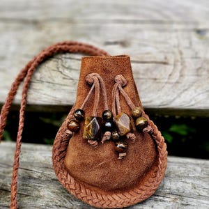 May include: A small, brown suede pouch with a braided trim and a braided strap. The pouch is adorned with decorative beads in shades of brown and black. The pouch is designed to be worn around the neck.