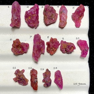 May include: A collection of fourteen raw, unpolished pink and red ruby crystals arranged in three rows on a white display tray. Each stone is numbered from 1 to 14, with a text label indicating 15-9mm, showcasing varied irregular shapes and textures.