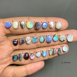 Puede incluir: Una colección de veintisiete ópalos pulidos sueltos de diversas formas y colores, incluyendo tonos azules, blancos e iridiscentes. Cada piedra está numerada del 1 al 27, con un texto que indica un tamaño de 9-7 mm para las piedras.